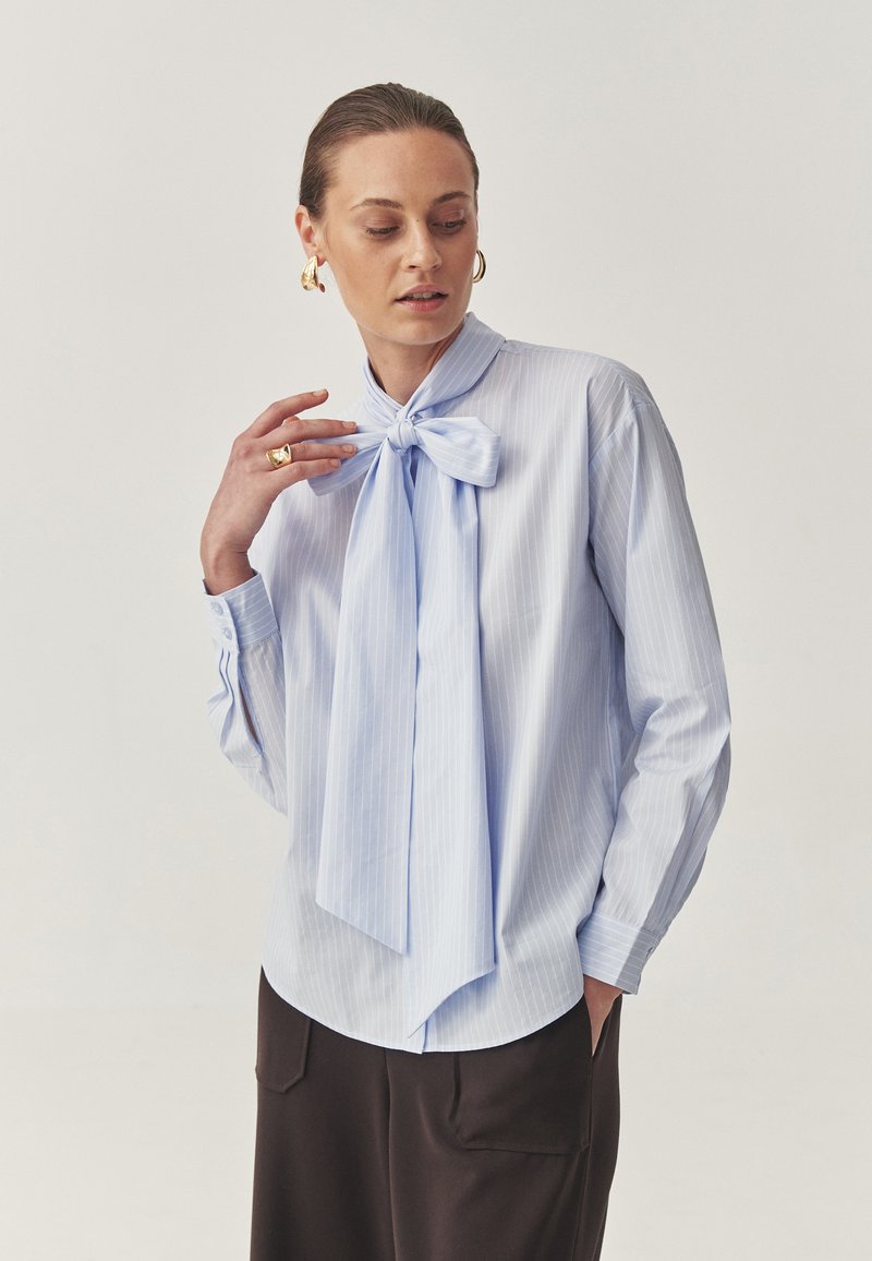Light blue striped shirt with a large bow tie at the neck, long sleeves, and button cuffs; smooth fabric with a relaxed fit.
