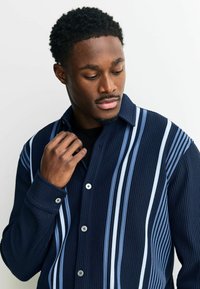 Mėlynos dryžuotos marškiniai su mygtukais, pasižymintys vertikaliais raštais skirtingų atspalvių mėlyna, su klasikinio tipo apykakle ir dviem priekiniais mygtukais.