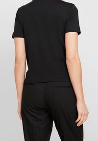 Osoba w gładkiej czarnej koszulce z krótkim rękawem i czarnych spodniach, pokazana z tyłu na jasnoszarym tle.