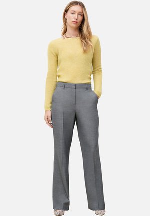Femme portant un pull jaune à manches longues, un pantalon gris sur mesure et des chaussures à motif peau de serpent, debout avec une main dans la poche.