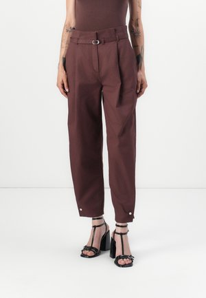 Personne portant un pantalon marron taille haute à coupe fuselée avec une passants de ceinture et des sandales noires à talons hauts avec bride en T, sur fond blanc.
