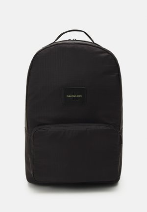 Zaino nero con una texture liscia, forma rotonda, tasca frontale con zip e una toppa del logo in verde. Realizzato in tessuto resistente con strisce sottili.