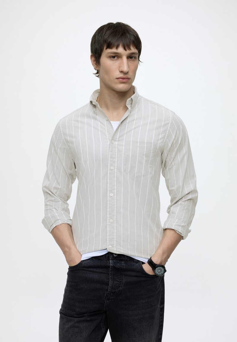 Ung mand med kort mørkt hår iført en lys beige stribet skjorte, sorte jeans og et sort armbåndsur, står med hænderne i lommerne.