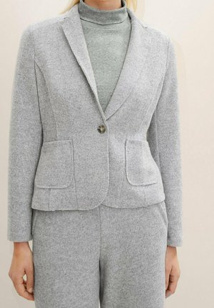 Vrouw draagt een lichtgrijze gebreide blazer over een bijpassende gebreide coltrui en broek, staand tegen een lichte neutrale achtergrond.