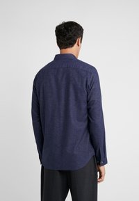 Man in een donkerblauw overhemd met lange mouwen en donkere broek, met zijn rug naar de camera gekeerd tegen een effen lichte achtergrond.