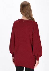 Burgunderfarbene Oversized-Strickjacke mit weitem Ausschnitt, überschnittenen Schultern und gerippten Bündchen. Weiche Stricktextur mit kleinem Logo auf der Rückseite.