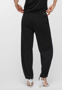 Person iført løstsiddende sorte bukser og sølvfarvede høje hæle-sandaler, justerer taljebåndet, set bagfra mod en enkel baggrund.