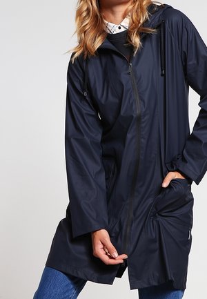 Personne portant un imperméable bleu marine avec une fermeture éclair et une capuche, tirant la partie avant du manteau vers le bas tout en se tenant devant un fond uni.