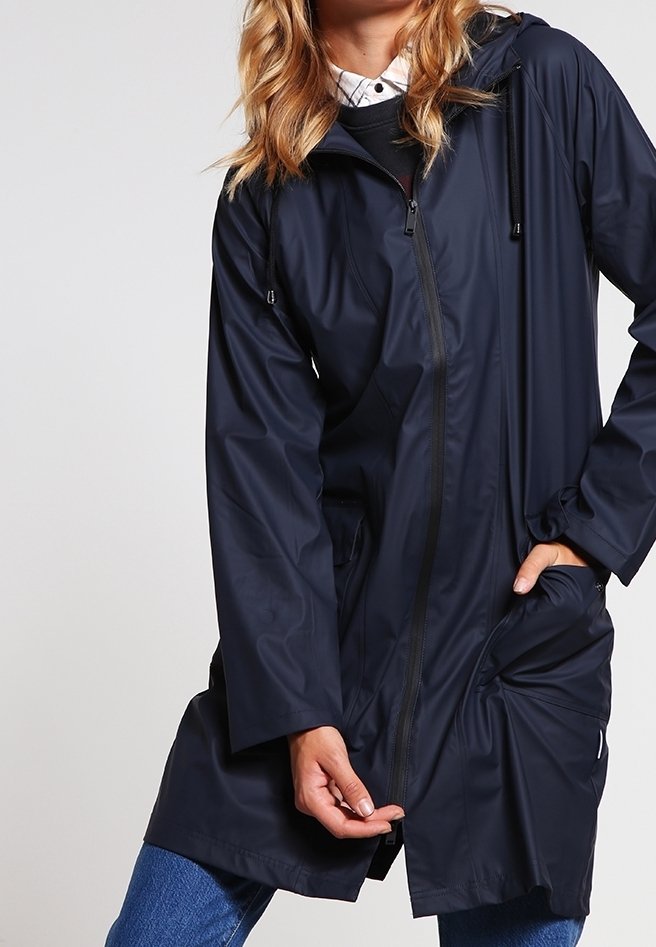 Personne portant un imperméable bleu marine avec une fermeture éclair et une capuche, tirant la partie avant du manteau vers le bas tout en se tenant devant un fond uni.