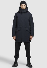 Parka marin cu guler înalt, multiple buzunare frontale și accente cu fermoar, combinat cu pantaloni negri și cizme negre cu șireturi.