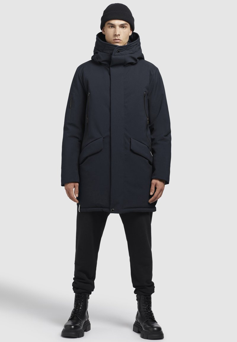 Parka marin cu guler înalt, multiple buzunare frontale și accente cu fermoar, combinat cu pantaloni negri și cizme negre cu șireturi.