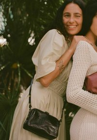 Svart handväska i krokodilprägling med rektangulär form och avtagbar axelrem. Två kvinnor bär ljusa och texturerade kläder.