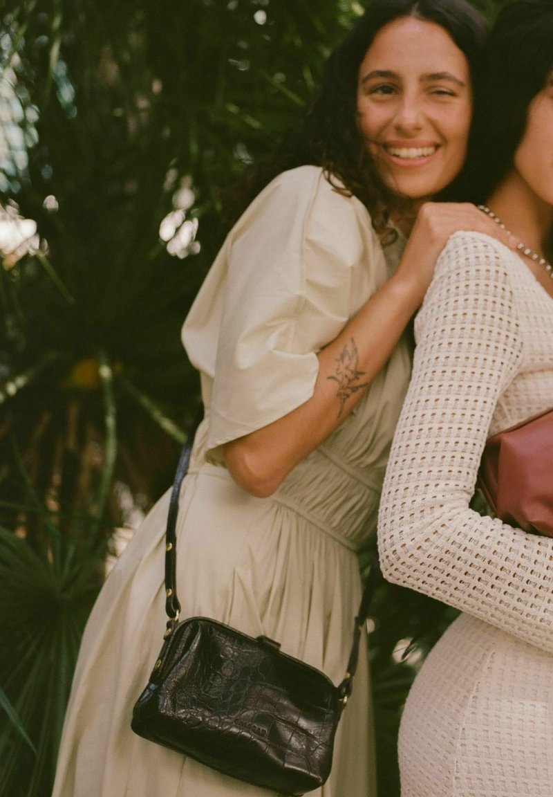 Svart handväska i krokodilprägling med rektangulär form och avtagbar axelrem. Två kvinnor bär ljusa och texturerade kläder.