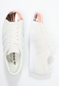 Valkoiset mokkanahkaiset tennarit, joissa on kolme teksturoitua raitaa, kiiltävä ruusukultainen kärki ja valkoinen kumipohja.