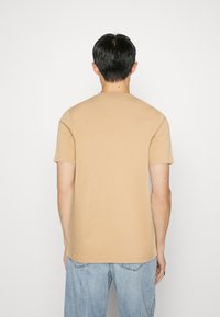 Beige színű, rövid ujjú pamut póló, kerek nyakkal és laza fazonnal. Texturált, sima anyag. Hátulról látható.