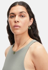 Groene geometrische oorbellen en een bijpassende ketting met gouden schakels, gedragen met een grijze mouwloze top, die een gladde textuur en gedefinieerde vormen heeft.