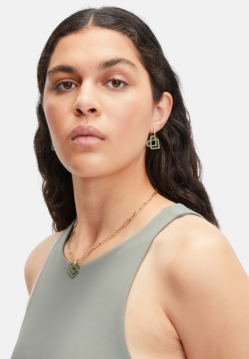 Groene geometrische oorbellen en een bijpassende ketting met gouden schakels, gedragen met een grijze mouwloze top, die een gladde textuur en gedefinieerde vormen heeft.