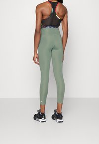 Leggings verdi oliva a vita alta, realizzati in tessuto elasticizzato, con una vestibilità aderente e un sottile marchio vicino alla caviglia. Piedi in scarpe da ginnastica nere.