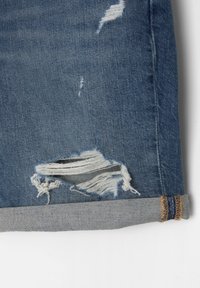 Tissu en denim bleu avec une déchirure effilochée au-dessus du revers roulé montrant des fils blancs et des détails de couture sur l'ourlet.