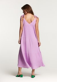 Rochie maxi dintr-un material texturat de nuanță lila, având bretele subțiri și un decolteu în V la spate, cu straturi suprapuse și o croială lejeră. Pantofi cu toc bloc verde.