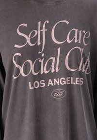 Grå langermet skjorte med et falmet rosa tekstdesign som inkluderer "Self Care," "Social Club," "Los Angeles," og "1995."