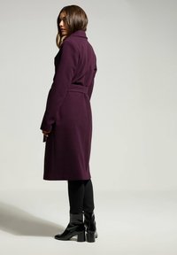 Vyötäröltä vyötetty, pitkä violetti takki, jossa on lovettui kaulus, villasekoitemateriaali ja sileä pinta, yhdistettynä mustiin slim-fit-housuihin ja kiiltäviin korokepolkimiin.