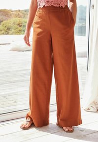 Pantalons larges en orange brûlé, fabriqués en tissu léger, avec une taille élastique et des poches latérales. Accompagnés de tongs.