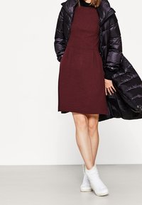 Burgundská pletená šaty se přiléhavým vrškem a rozšířenou sukní, doplněné černou zateplenou bundou. Modelka má na sobě bílé vysoké tenisky.