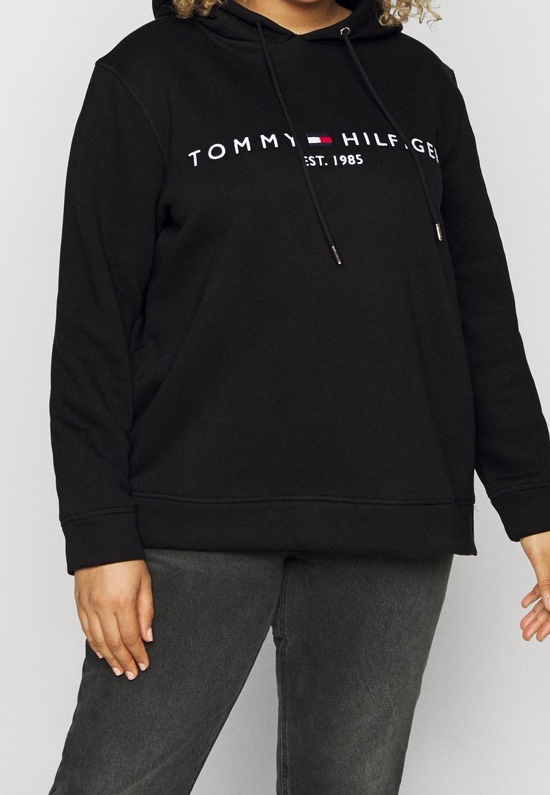 Personne portant un sweat à capuche noir avec le logo "Tommy Hilfiger Est. 1985" et un pantalon gris foncé, debout devant un fond clair uni.