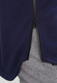 Marinblå zip-up topp med en mjuk tygtextur, som har en svart dragkedja. Bärs över ett grått plagg och visar ett lagerdesign.