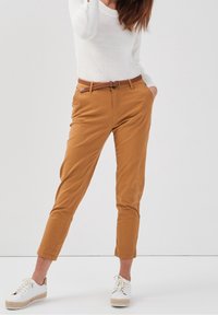 Chino en coton beige avec une coupe droite, featuring une ceinture en cuir tressé marron, des poignets retroussés, et associés à des espadrilles blanches.