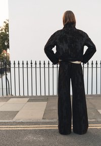 Jachetă din catifea brună și pantaloni largi, cu o suprafață texturată și croială scurtă. Vedere din spate, cu un zid alb simplu în fundal.