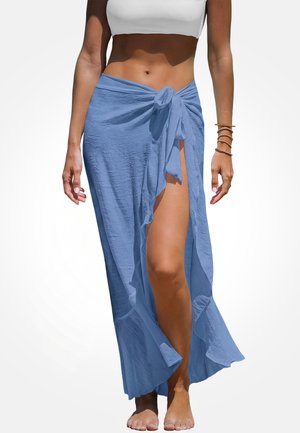 Vrouw draagt een lichtblauwe wikkelrok met een voorste split, gestrikt in de taille, gecombineerd met een witte cropped top en gouden armbanden.