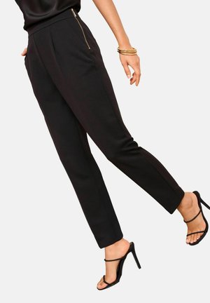 Femme portant un pantalon noir fuselé ajusté avec une fermeture éclair latérale, associé à des sandales noires à talons hauts et des bracelets dorés à un poignet.