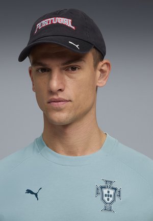 Man met een zwarte pet met de tekst "PORTUGAL" en een lichtblauw sportshirt met Puma-logo en het voetbalwapen van Portugal.