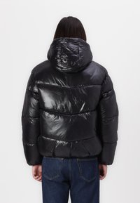 Sort pufferjakke med en skinnende tekstur, hætte og vandret quiltet design. Sammen med blå jeans, set fra ryggen.
