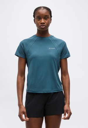 Γυναίκα που φοράει μια τιρκουάζ κοντομάνικη μπλούζα Columbia και μαύρο σορτς, στέκεται με πρόσωπο προς τα εμπρός μπροστά σε ένα απλό λευκό φόντο.