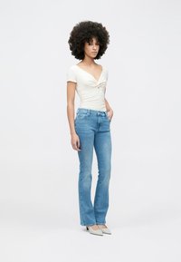 White fitted top with a twist detail, paired with light blue flared jeans. The outfit includes light gray pointed shoes. Solid color background.
