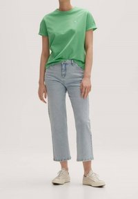 Groene katoenen t-shirt met opgerolde mouwen en klein hartlogo, gecombineerd met lichtblauwe rechte jeans en beige sneakers.