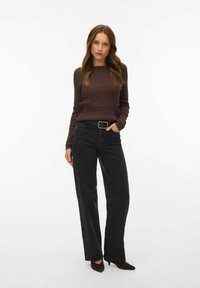 Femme debout, une main dans la poche, portant un pull marron en maille torsadée, un jean noir taille haute avec ceinture, et des talons pointus foncés.