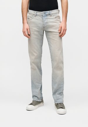 Man draagt lichtgewassen, slim-fit jeans met lichte slijtage, gecombineerd met olijfgroene sneakers met witte zolen en veters.