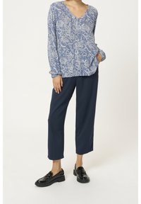 Blaue gemusterte Bluse mit langen Ärmeln und V-Ausschnitt, kombiniert mit marineblauen weiten Hosen und schwarzen glänzenden Loafers.