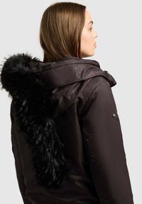Dunkelbraune Steppjacke mit einer gefütterten Kapuze aus Fell, glatter Oberfläche, Druckknopfverschluss und einem seitlichen Knopfdetail nahe der Schulter.