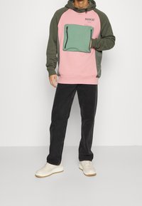 Person som bär en rosa och grön Nike-hoodie med framficka, svarta byxor och beige sneakers stående mot en vit bakgrund.