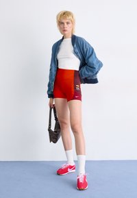 Denim bomber jakna preko bijele majice, uparena s crvenim i tamnim šortsem s Nike logom. Ružičaste tenisice s bijelim čarapama, drži smeđu torbu.