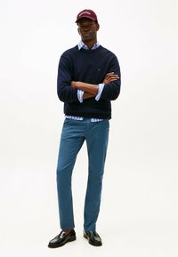 Maglione di maglia blu navy sopra una camicia azzurra a righe con bottoni, pantaloni turchesi e mocassini neri. Un berretto bordeaux con testo bianco completa l'outfit.