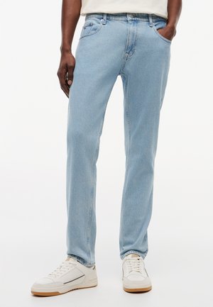 Άνδρας που φοράει ανοιχτό μπλε τζιν παντελόνι και λευκά αθλητικά παπούτσια, στέκεται με το ένα χέρι στην τσέπη μπροστά σε απλό φόντο.