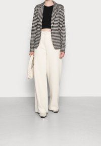 Czarno-biała marynarka w kratę, czarny krótki top, kremowe szerokie spodnie, trzymająca kremową plecioną torebkę i nosząca beżowe buty z metalicznymi czubkami.