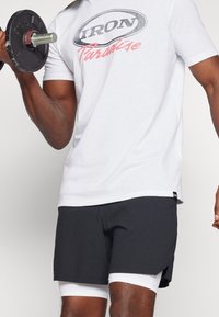 Biała koszulka z nadrukiem "IRON Paradise" w odcieniach szarości i różu, w zestawie z ciemnymi szortami z białym podszewką i bocznymi rozcięciami.