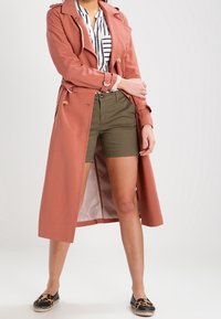 Donna con un lungo trench coat color ruggine, camicia a righe, pantaloni corti verdi oliva e espadrillas a stampa leopardata, in piedi con le braccia incrociate.
